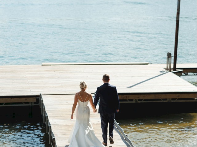 A couple in wedding attire is walking hand in hand down a wooden pier towards the water on a sunny day.