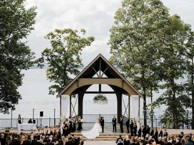 A wedding ceremony by a lakeside under a decorated wooden pavilion, surrounded by guests in formal attire.