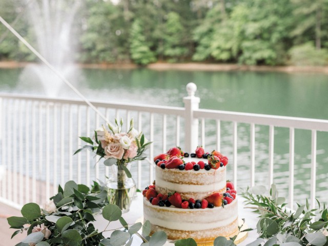 A two-tiered naked cake adorned with berries on a decorated table, overlooking a pond with a fountain and lush greenery in the background.