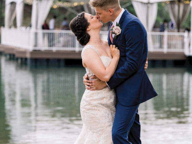 A couple in wedding attire kisses by the water, with a pavilion in the background, all in a picturesque setting.