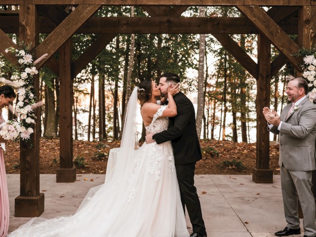 A bride and groom kiss under a wooden arch in an outdoor wedding ceremony, surrounded by floral arrangements and applauding guests.