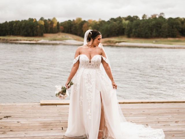 A bride in a white wedding dress stands on a wooden dock by a lake, holding a bouquet, with trees visible in the background.