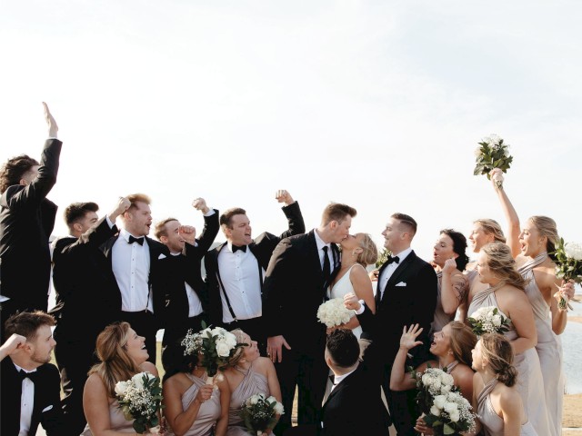 A joyful bridal party celebrates a wedding, with the couple kissing amidst cheers and friends holding flowers.