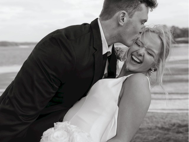 A smiling couple embraces outdoors; one kisses the other on the forehead, holding a bouquet of roses.