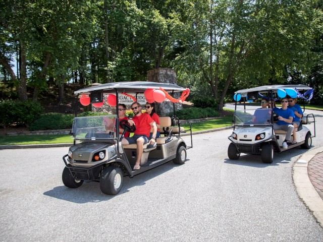 People in decorated golf carts drive on a sunny day, festooned with red and blue balloons and ribbons, surrounded by trees.