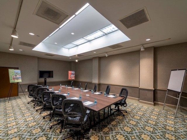 A conference room with a rectangular table, chairs, a flip chart, a flat-screen TV, water bottles, paper, and pens under a skylight ceiling.