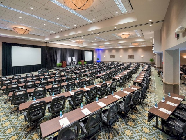 The image shows a large conference room set up with rows of tables and chairs facing two projection screens and a stage at the front.