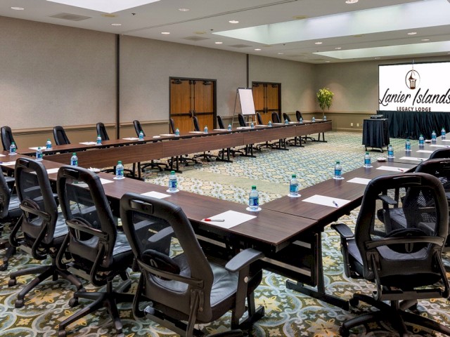 A large conference room with a U-shaped table setup, chairs, notepads, water bottles, and a projector displaying 