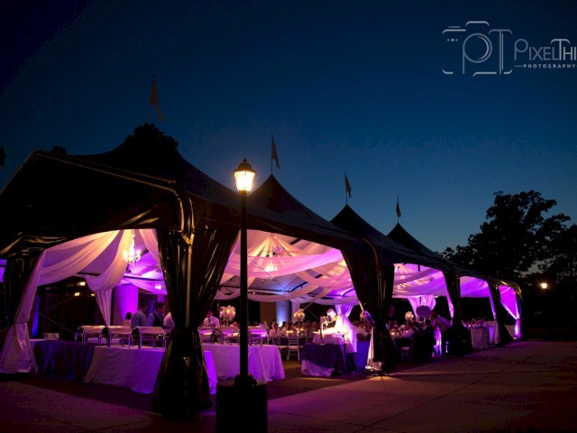 The image shows an outdoor event tent draped in fabric with purple lighting, set up in the evening with tables and chairs arranged inside.