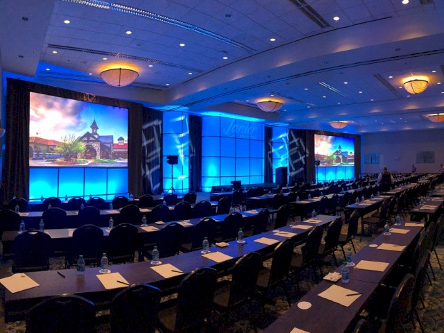 The image shows a large conference room set up for an event, featuring rows of tables and chairs, two large screens, and blue lighting throughout the room.