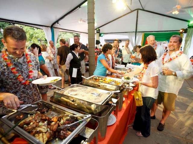 People at a party buffet in a tent, serving themselves from various trays of food, and socializing, with some wearing leis around their necks.