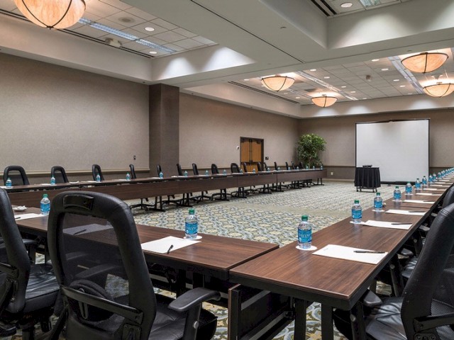 A large conference room setup with U-shaped tables, chairs, water bottles, notepads, pens, and a projector screen, ready for a meeting.
