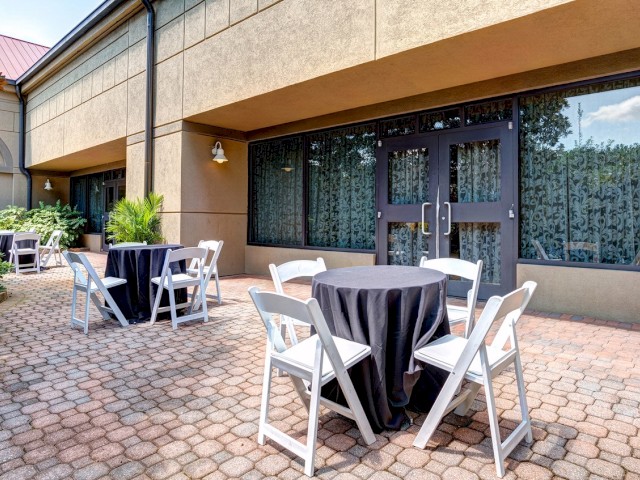 The image shows an outdoor seating area with tables covered in black tablecloths, surrounded by white chairs, and set on a brick-paved patio.