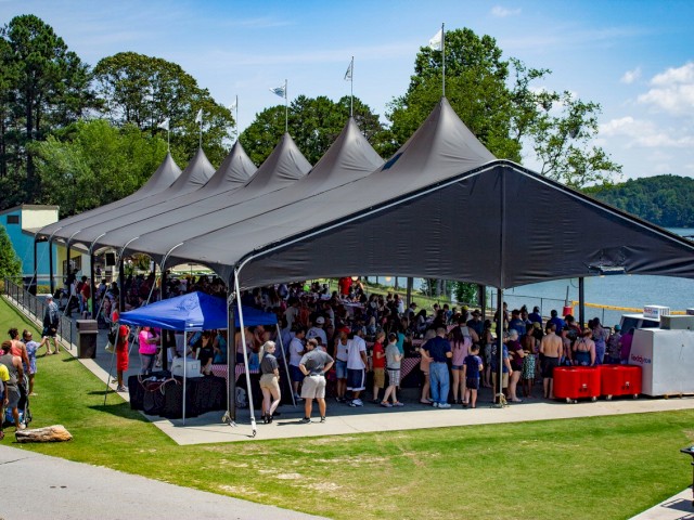 The image shows a large outdoor event with people gathered under a large tent near a body of water, with food and drink stands around.