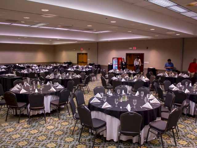 A large banquet hall is set up with round tables covered in black and white linens, with neatly arranged place settings and glasses.
