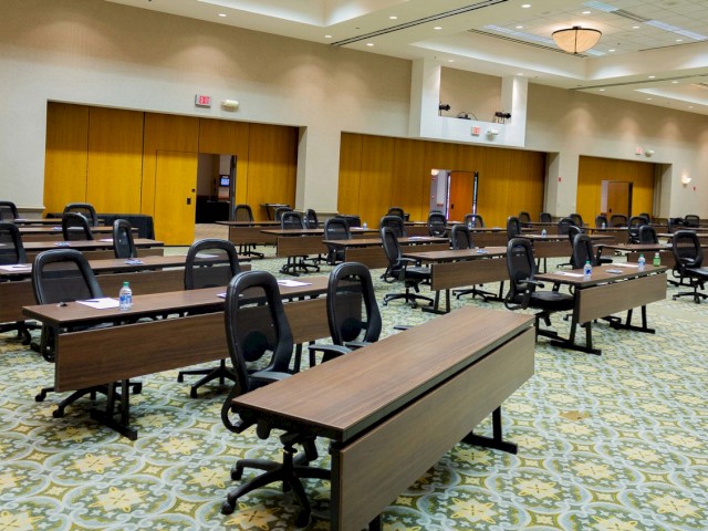 The image shows an empty conference room with rows of tables and office chairs, a patterned carpet, and a few water bottles on the tables.