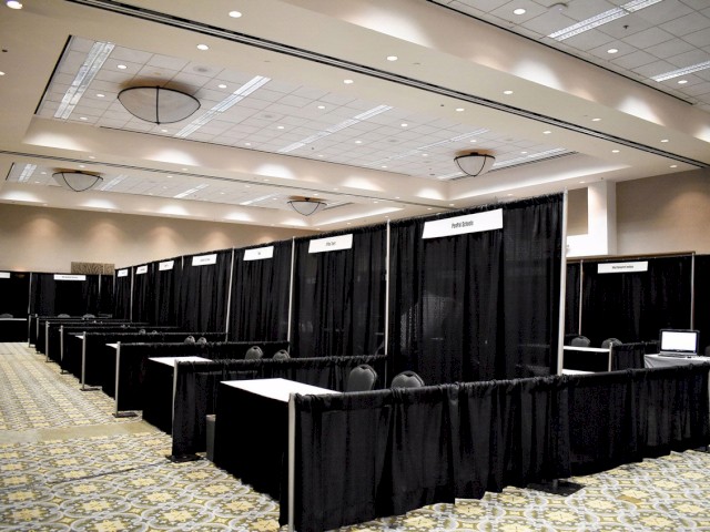 An empty convention hall with booths set up, each separated by black curtains and labeled with different signs, ready for an event to take place.