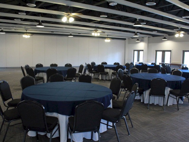 The image shows a banquet hall with round tables covered in blue tablecloths and surrounded by black chairs, well-lit with ceiling lights.