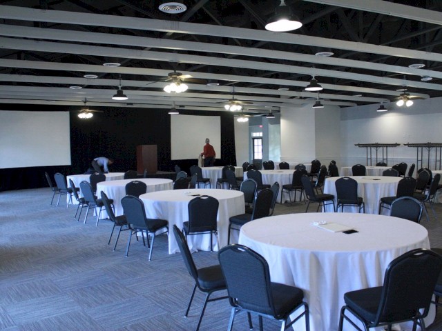 A conference room setup with round tables covered in white cloth, black chairs, projection screens, and a few people in view.