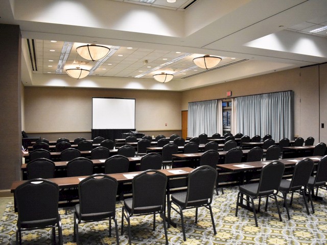 An empty conference room with rows of tables and chairs, a projector screen at the front, and ceiling lights illuminating the space.