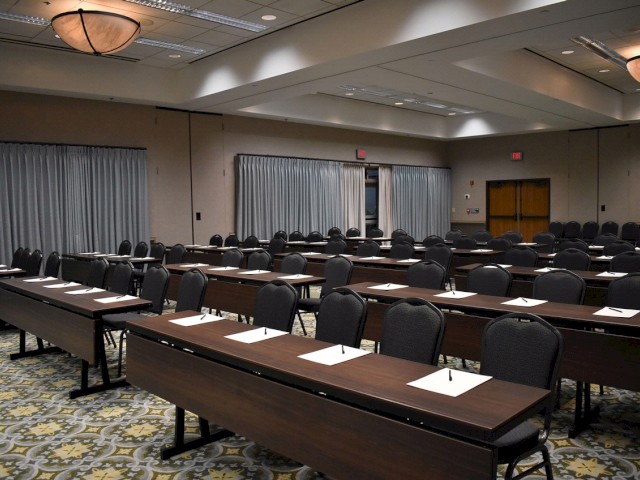 A conference room with rows of tables and chairs, each set with a notepad and pen. The room has curtains, carpeted flooring, and overhead lighting.