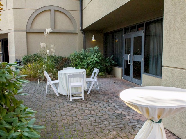 An outdoor patio with a table with white chairs, another covered table, greenery, brick flooring, and double glass doors leading to the building.