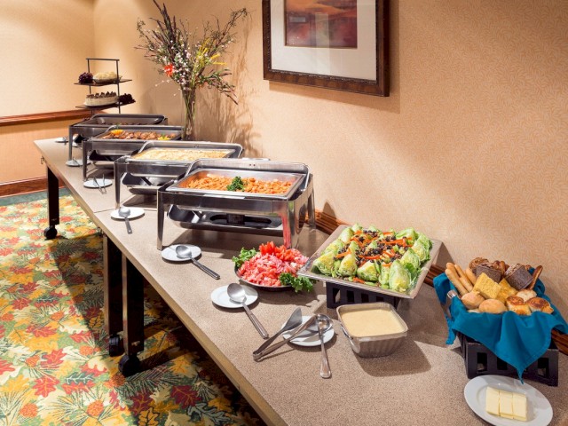 The image shows a buffet setup with chafing dishes, plates of salad, bread, sliced vegetables, and sauces on a long table in a decorated room.