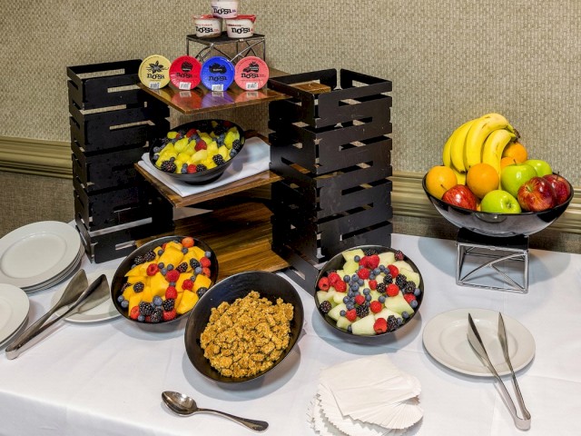 A breakfast buffet displaying yogurt, assorted fruits, granola, and fresh fruit bowls set on a white tablecloth with cutlery and plates.