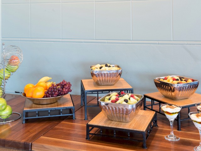 An assortment of fresh fruit bowls, parfaits, and whole fruits displayed on a wooden and metal stand on a countertop, ready for serving.