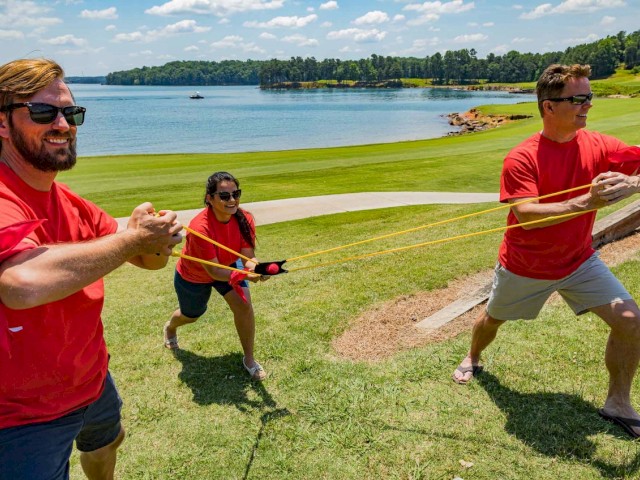 Three people wearing red shirts are outdoors by a lake, playing with a large slingshot, stretching it while standing on grass.