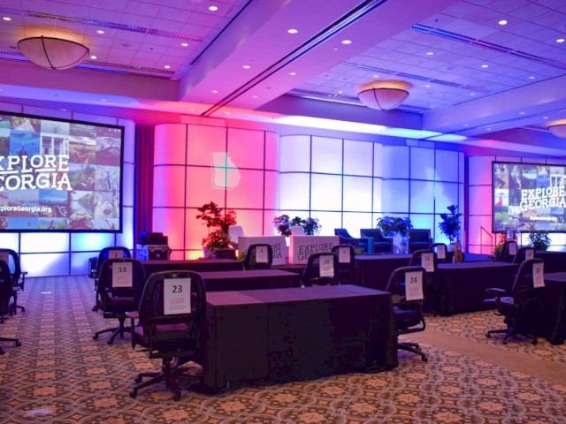 The image shows a conference room setup with desks and chairs, two screens displaying 