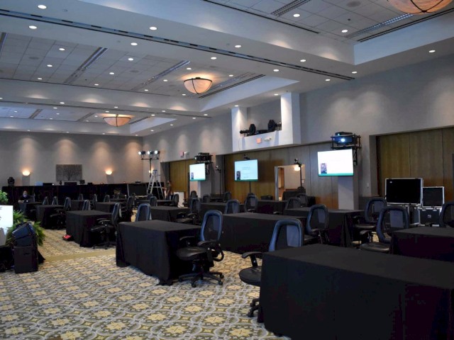 The image shows a conference room with black tables and chairs, multiple screens, and lighting equipment, with a patterned carpet and ceiling lights.