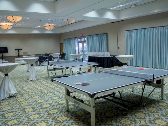 A room with ping pong tables, cocktail tables, and chairs arranged for a social or recreational event, with blue curtains and patterned carpet.