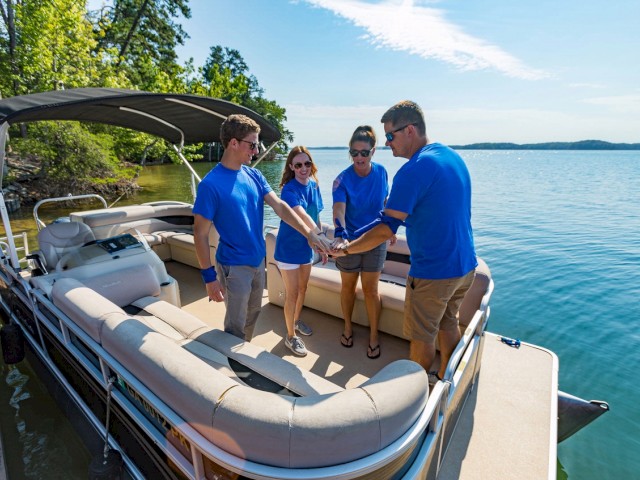 Four people in blue shirts stand on a boat, joining hands in a circle, with a scenic lake and trees in the background.