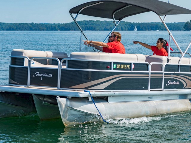 Two people on a pontoon boat are pointing towards something on a lake with calm waters and forested shores in the background.
