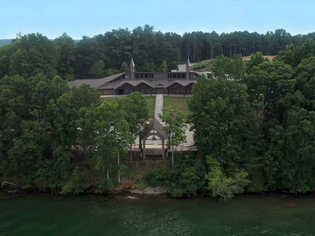 An aerial view of a large wooden building complex surrounded by dense trees, situated near a body of water. The structures are nestled in a forested area.
