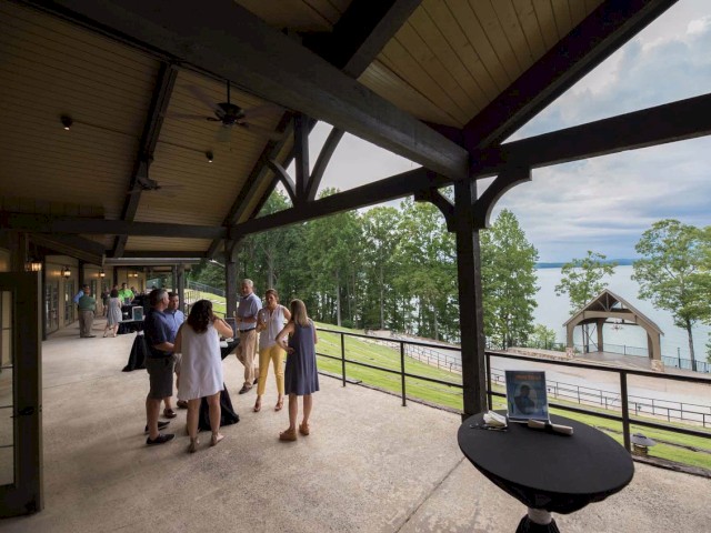 People are gathered under a covered patio near a scenic lake, conversing and gesturing. A round table holds a picture frame on the side.
