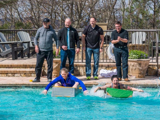 People standing poolside watching as two individuals race in the water with cardboard boxes.