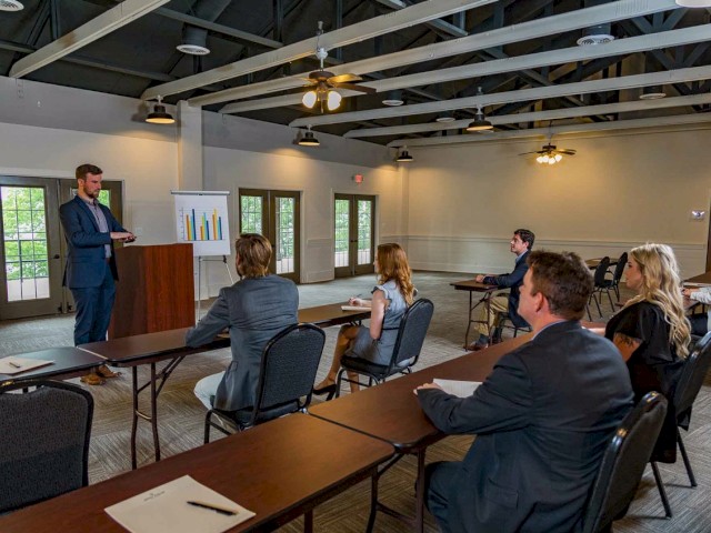 A man is giving a presentation to a small group of people seated at tables in a conference room, with a chart displayed beside him.