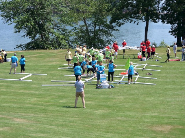 A group of people, divided into teams by their shirt colors, is assembling structures on a grassy area near a body of water, with trees in the background.