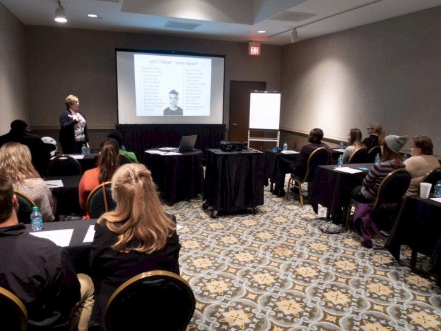 A group of people attends a presentation in a conference room, with the speaker standing in front of a projector screen and whiteboard, facing the audience.