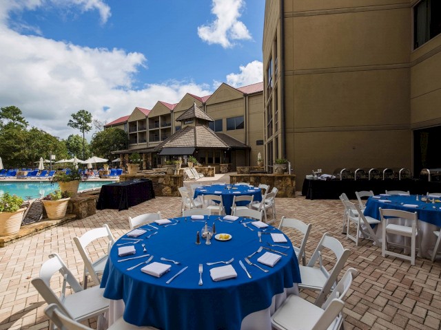 Outdoor dining setup with blue tablecloths and white chairs near a poolside at a resort, featuring a stone building and a partly cloudy sky.