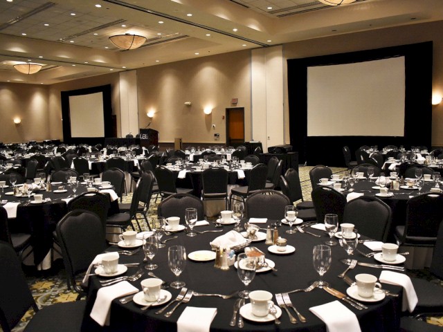 The image shows a banquet hall set up with round tables, black tablecloths, and white napkins, with formal place settings and two large projection screens.