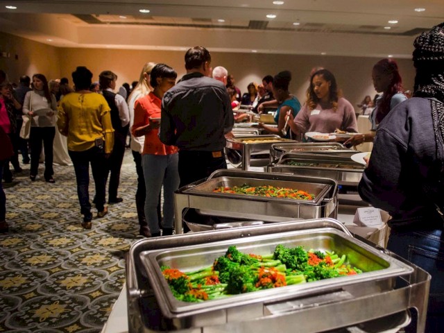 People are gathering and serving food from buffet trays filled with vegetables at an indoor event, engaging and conversing with one another.