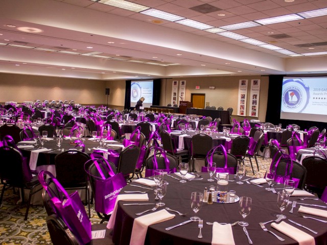 A conference room is set up for an event, with round tables, purple bags on chairs, and presentation screens at the front. The room is empty.