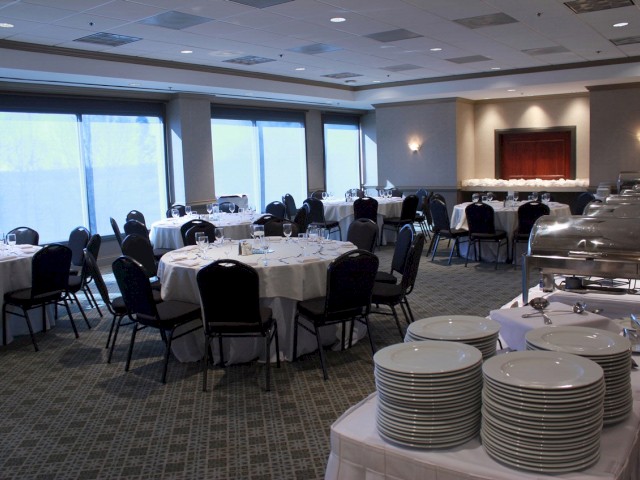 An empty banquet hall with round tables and chairs set up, buffet chafing dishes, and stacks of plates ready for an event, with large windows.