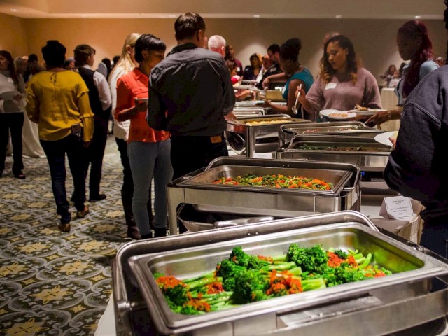 People are attending a buffet-style event with various dishes including vegetables, and some individuals are gathering food and socializing.