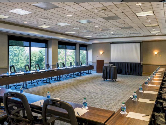 A conference room with a U-shaped arrangement of tables, water bottles, notepads, chairs, a projector screen, podium, and large windows is shown.