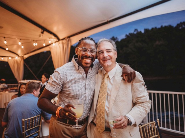Two men are smiling and posing together under a canopy with string lights. One has his arm around the other, both holding drinks.
