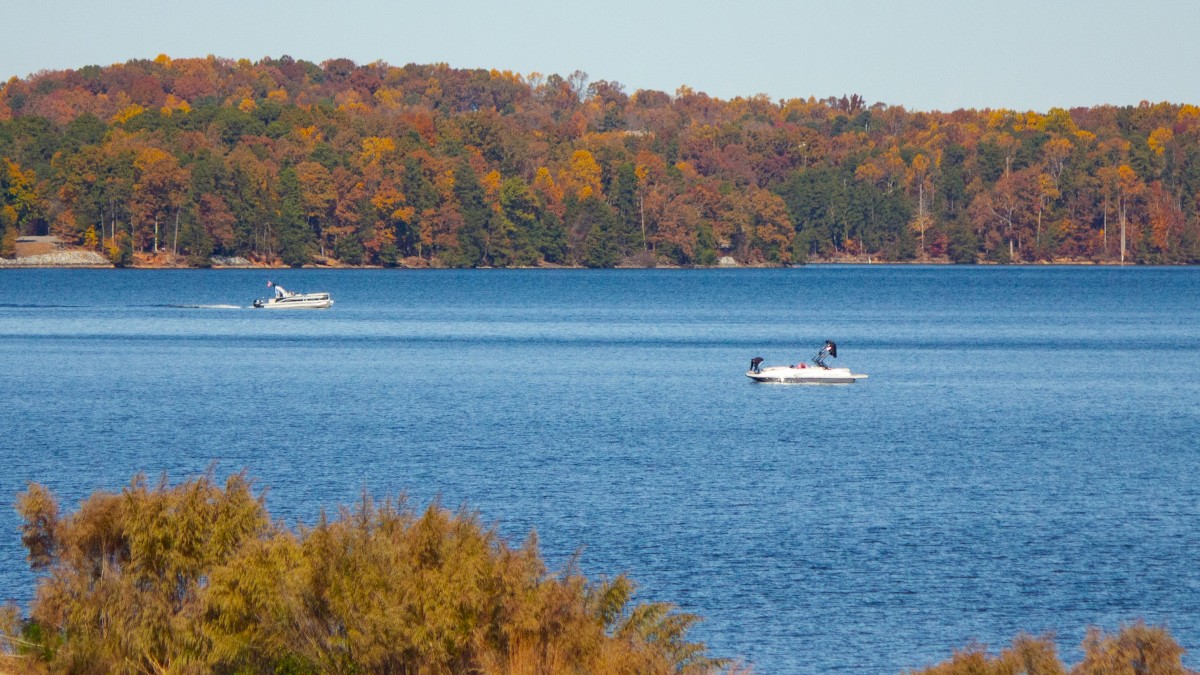 A tranquil lake with two boats, surrounded by autumn trees and grassy foreground under a clear sky.